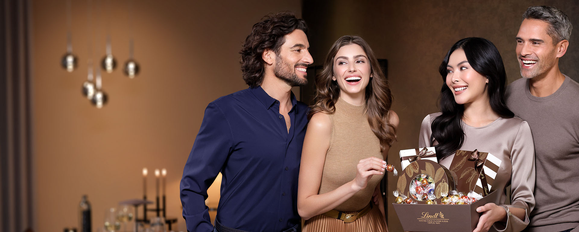 Two smiling couples, each including a woman and a man, standing in front of a laid table; one of the women is holding a Lindt present box in her hands (Photo)