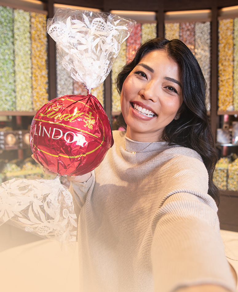 Eine braunhaarige, lächelnde Frau in einem Lindt-Shop mit einer großen roten LINDOR-Kugel in den Händen; im Hintergrund sind Behälter mit verschiedenen LINDOR-Kugeln (Foto)